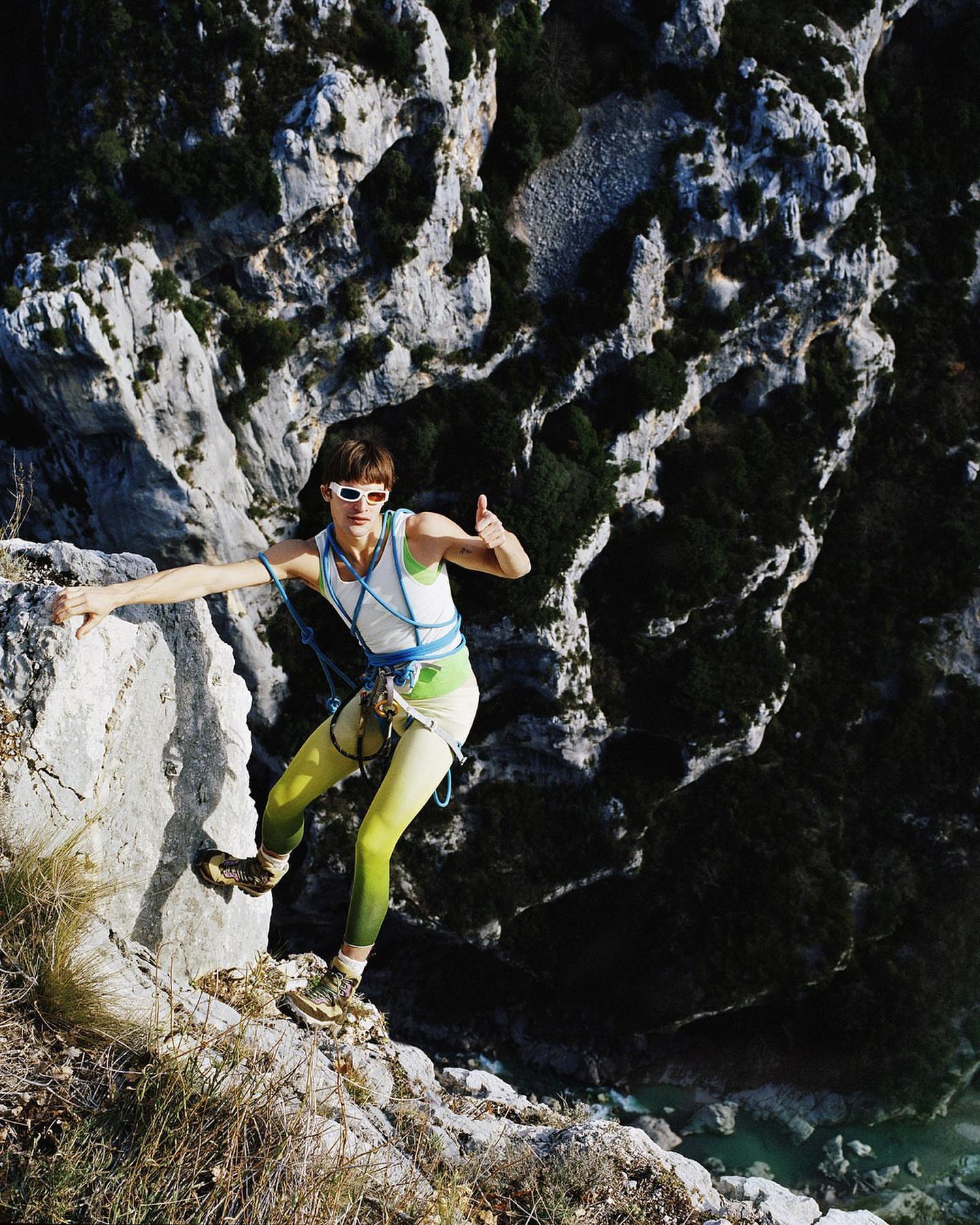 A woman rock climbing outdoors in yellow leggings and a white tank top, with a thumbs up, set against a rocky cliffside backdrop.