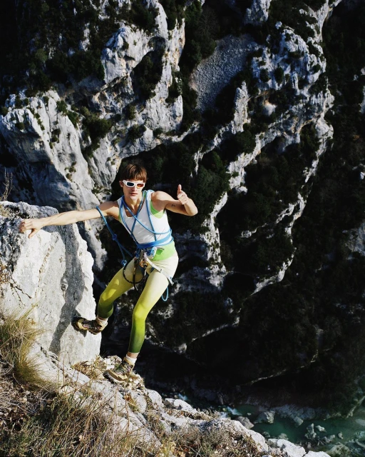 A woman rock climbing outdoors in yellow leggings and a white tank top, with a thumbs up, set against a rocky cliffside backdrop.