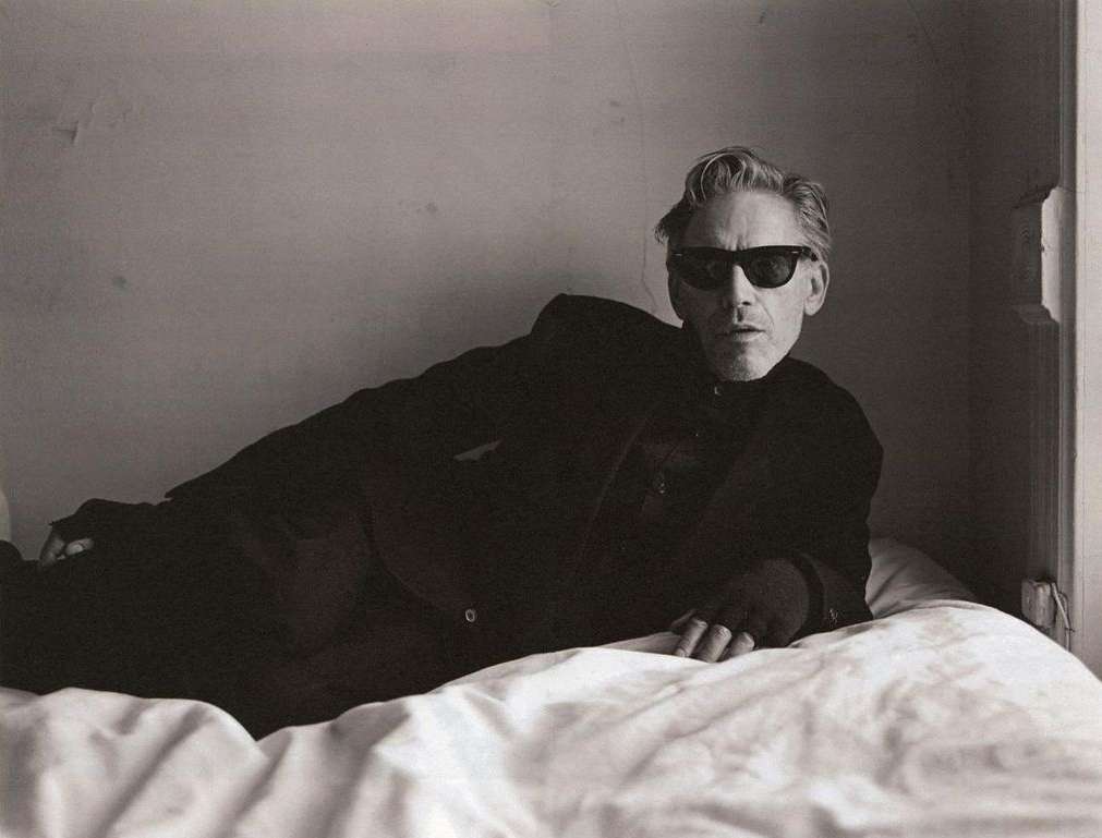 Man wearing dark sunglasses and a black suit reclining on a white bed in a minimalist room by photographer Brett Lloyd.