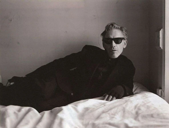 Man wearing dark sunglasses and a black suit reclining on a white bed in a minimalist room by photographer Brett Lloyd.