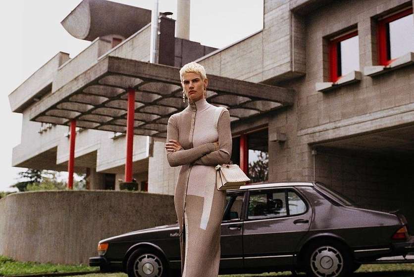 A fashion shot of a woman with short blonde hair wearing a neutral-toned dress, standing in front of a modern building and a dark car.