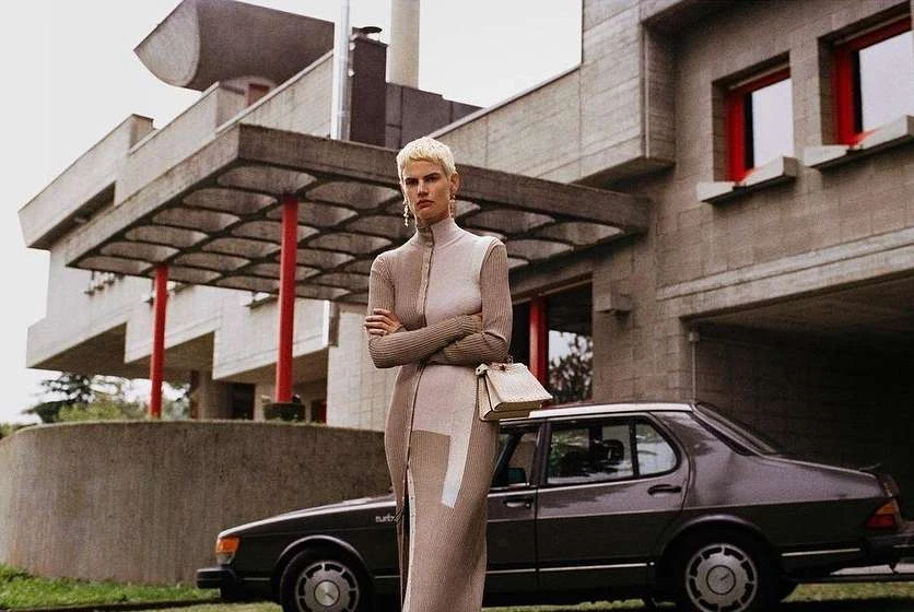 A fashion shot of a woman with short blonde hair wearing a neutral-toned dress, standing in front of a modern building and a dark car.