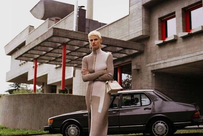 A fashion shot of a woman with short blonde hair wearing a neutral-toned dress, standing in front of a modern building and a dark car.