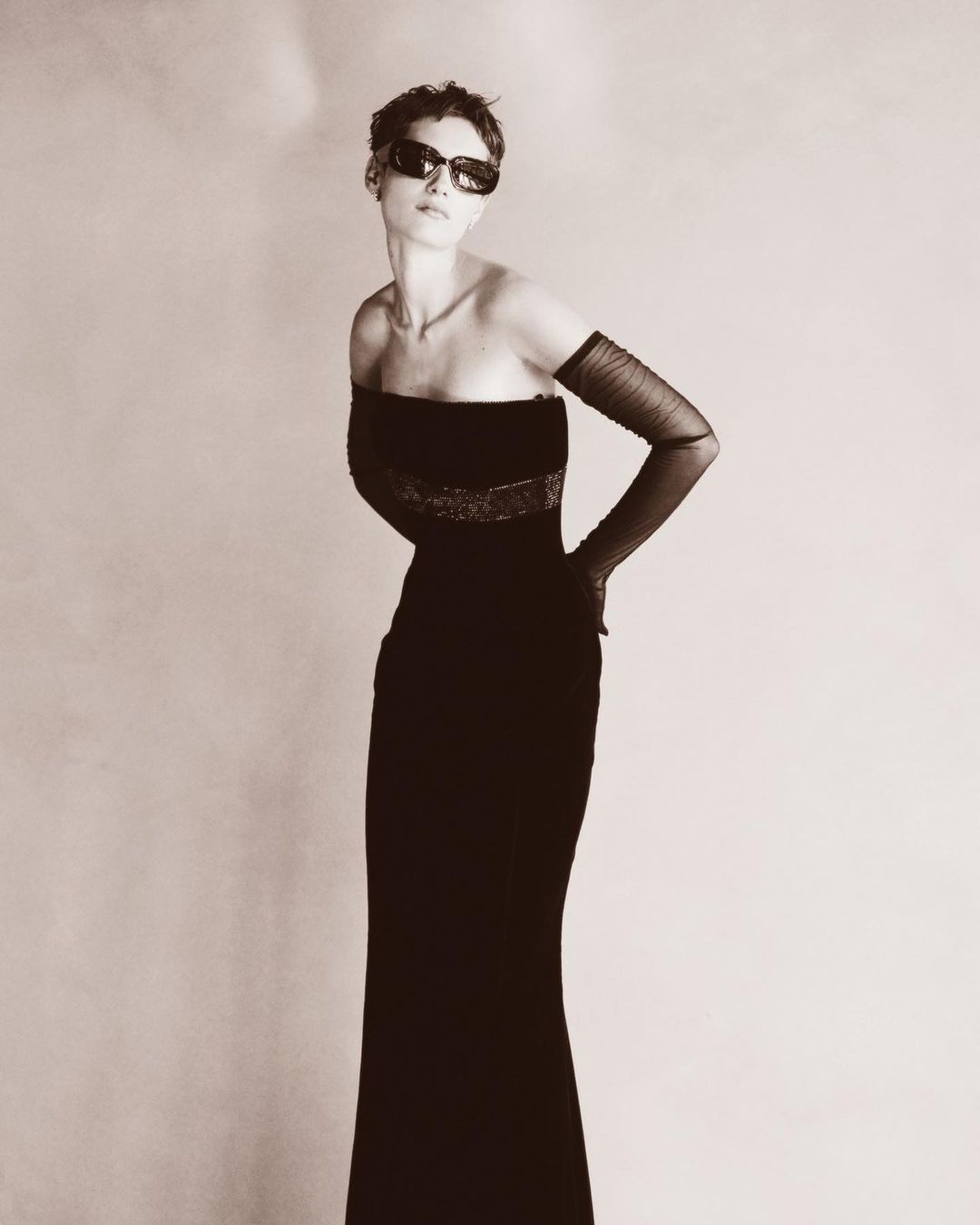 Model with short hair wearing sunglasses and a black strapless evening gown with sheer opera gloves