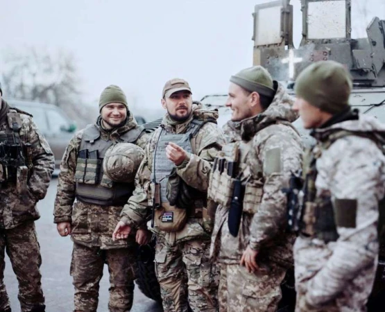 Group of soldiers in camouflage uniforms and beanies standing outdoors near military vehicles on a cloudy day, looking alert.