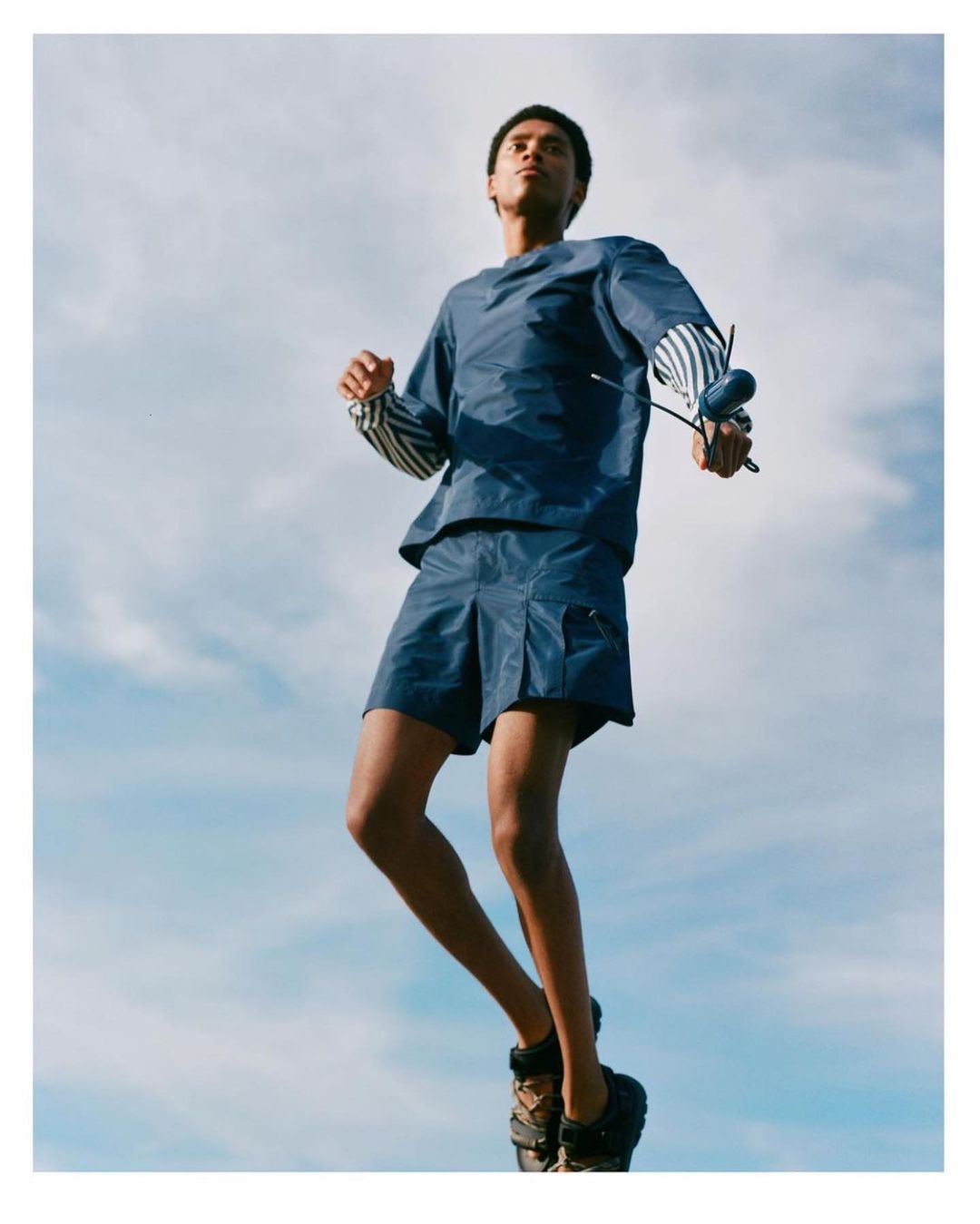 Fashion shot of a young adult man in blue shorts and shirt jumping against a blue sky background, showcasing a modern, athletic style.