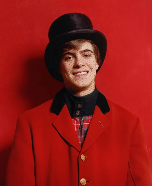 Smiling person in a red jacket, black top hat, and a black net in front of their face on a red background in a portrait orientation.