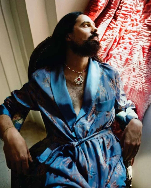 Man in patterned robe sitting in chair with draped red and white fabric in background; wearing necklace with jeweled pendant.  