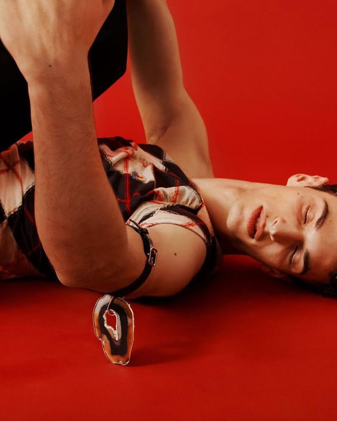 Young adult laying on a red surface with a large crystal pendant. The model is wearing a white and red patterned top and black suspenders.