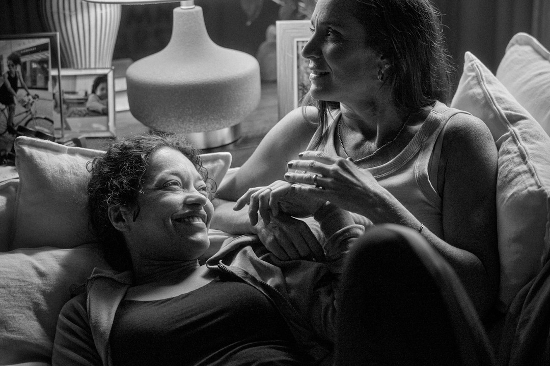 Monochrome image of two women lying on a couch, smiling and holding hands in an indoor setting with pillows and a lamp visible.