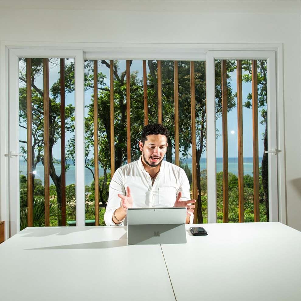 Adult male interacts with a tablet at a bright white table in a well-lit room, with trees and ocean visible through a window.