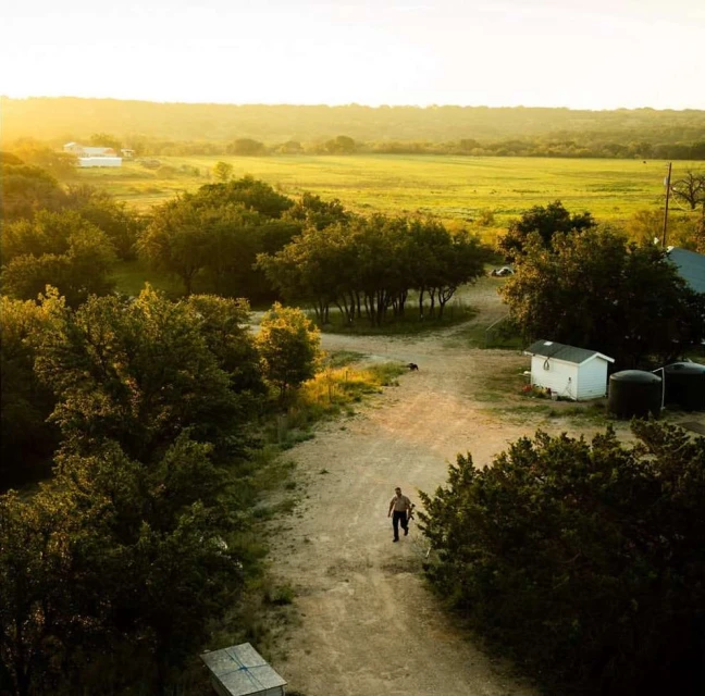 Aerial view of an adult male walking on a dirt road with green fields and scattered trees in the background during golden hour.