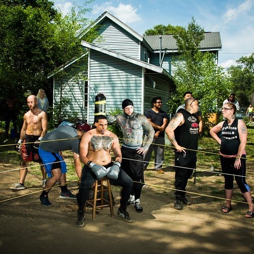 Outdoor boxing match with tattooed fighter seated in ring, surrounded by spectators, set against a backdrop of trees and a light blue house.