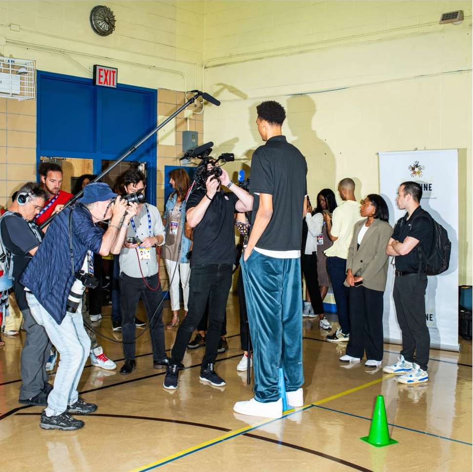 Tall basketball player poses surrounded by photographers and media personnel indoors in a gymnasium-like setting with bright lighting.