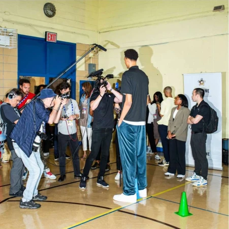 Tall basketball player poses surrounded by photographers and media personnel indoors in a gymnasium-like setting with bright lighting.