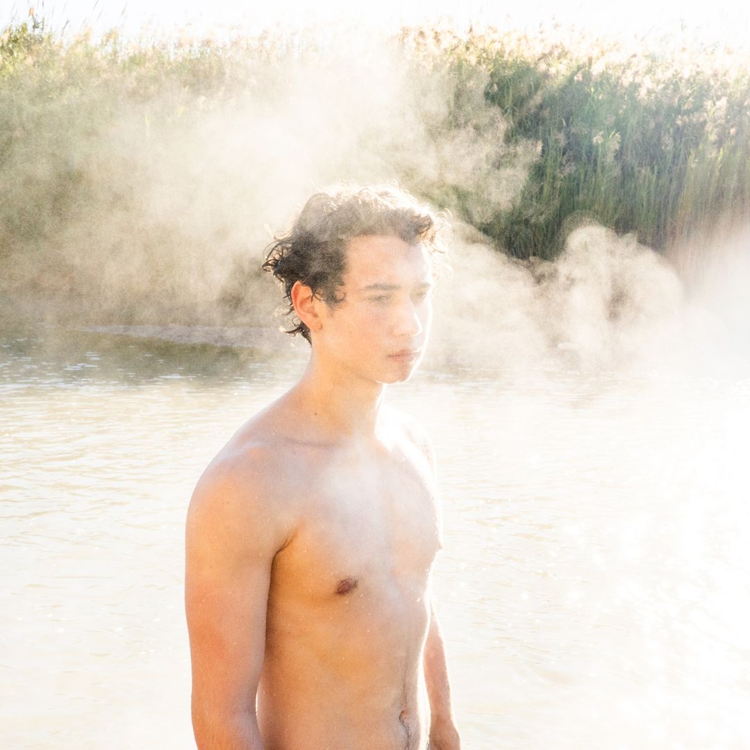 Shirtless young man stands in water with fog; reeds are visible in the background, bathed in soft, natural lighting.