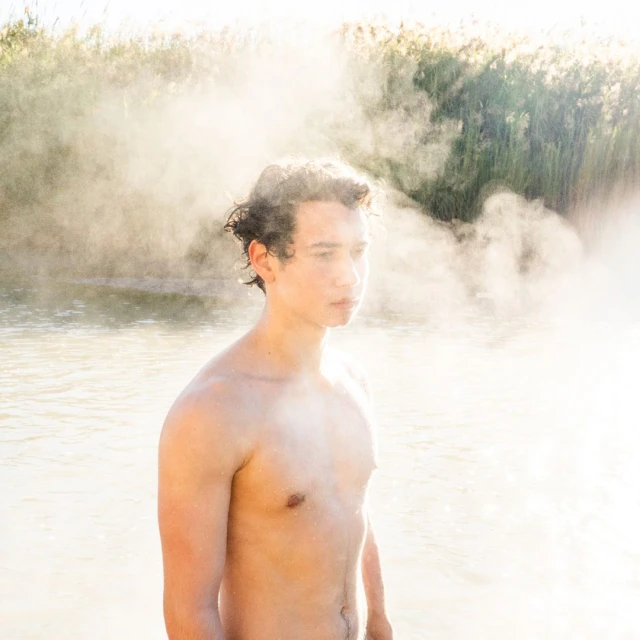 Shirtless young man stands in water with fog; reeds are visible in the background, bathed in soft, natural lighting.