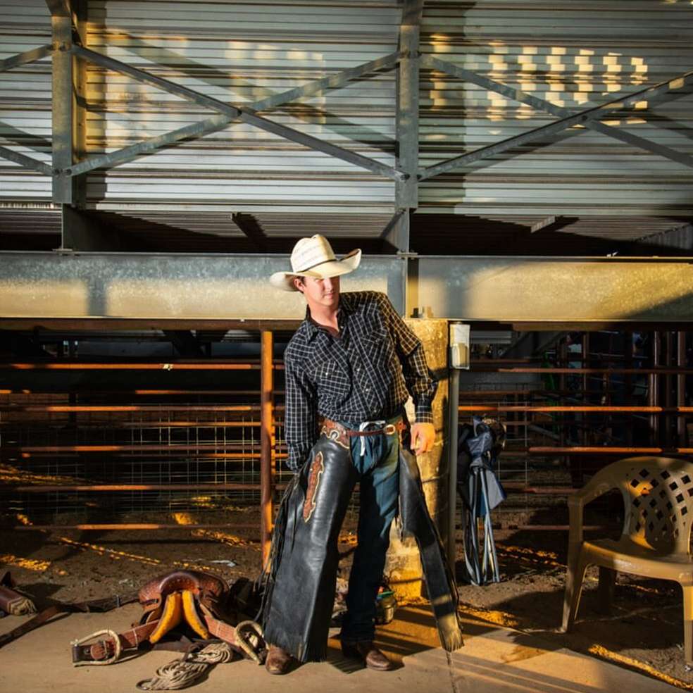 Cowboy standing in indoor arena wearing chaps and hat with saddle and chair in background, captured in medium shot.