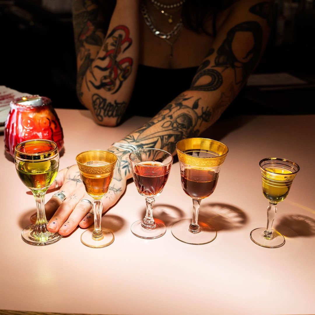 Close-up on a pink table with five stemmed glasses filled with colorful liquids, illuminated by a warm light, a tattooed arm visible in the background.