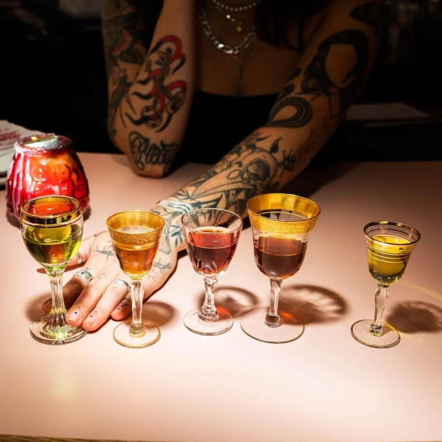 Close-up on a pink table with five stemmed glasses filled with colorful liquids, illuminated by a warm light, a tattooed arm visible in the background.