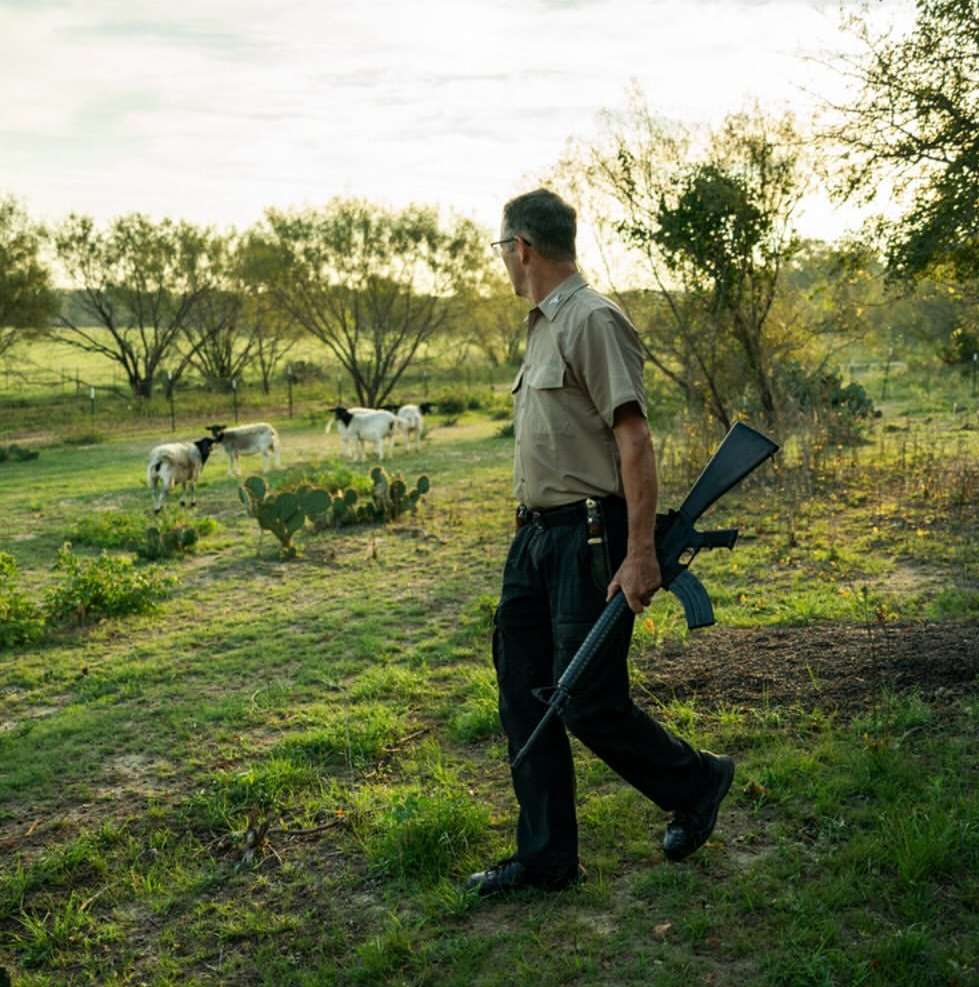 Man in khaki uniform carries rifle across grassy field, sheep graze in background, rural setting, outdoor daylight scene, natural lighting.