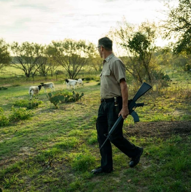 Man in khaki uniform carries rifle across grassy field, sheep graze in background, rural setting, outdoor daylight scene, natural lighting.