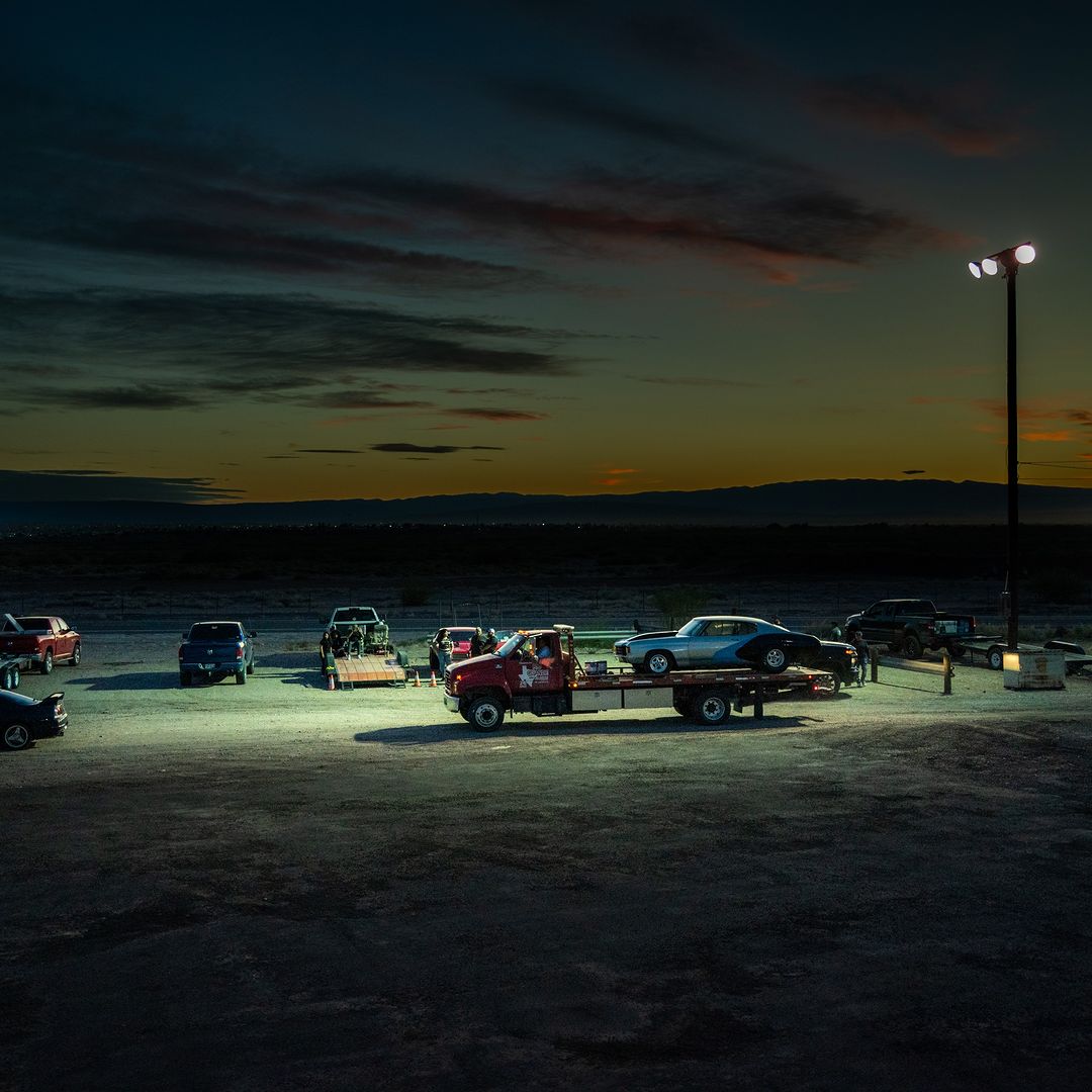At twilight, a flatbed truck carrying a blue classic car is parked in a lot, surrounded by other vehicles, under bright pole lights.