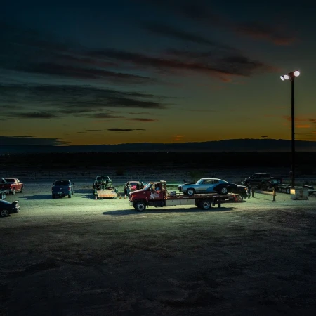 At twilight, a flatbed truck carrying a blue classic car is parked in a lot, surrounded by other vehicles, under bright pole lights.