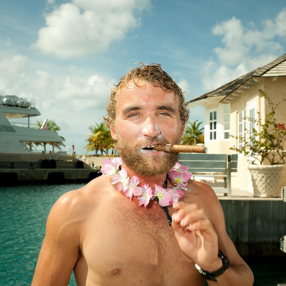 Adult man with wet hair holds a cigar in his mouth while wearing a flower lei necklace outside near a building and yacht.