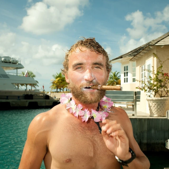 Adult man with wet hair holds a cigar in his mouth while wearing a flower lei necklace outside near a building and yacht.