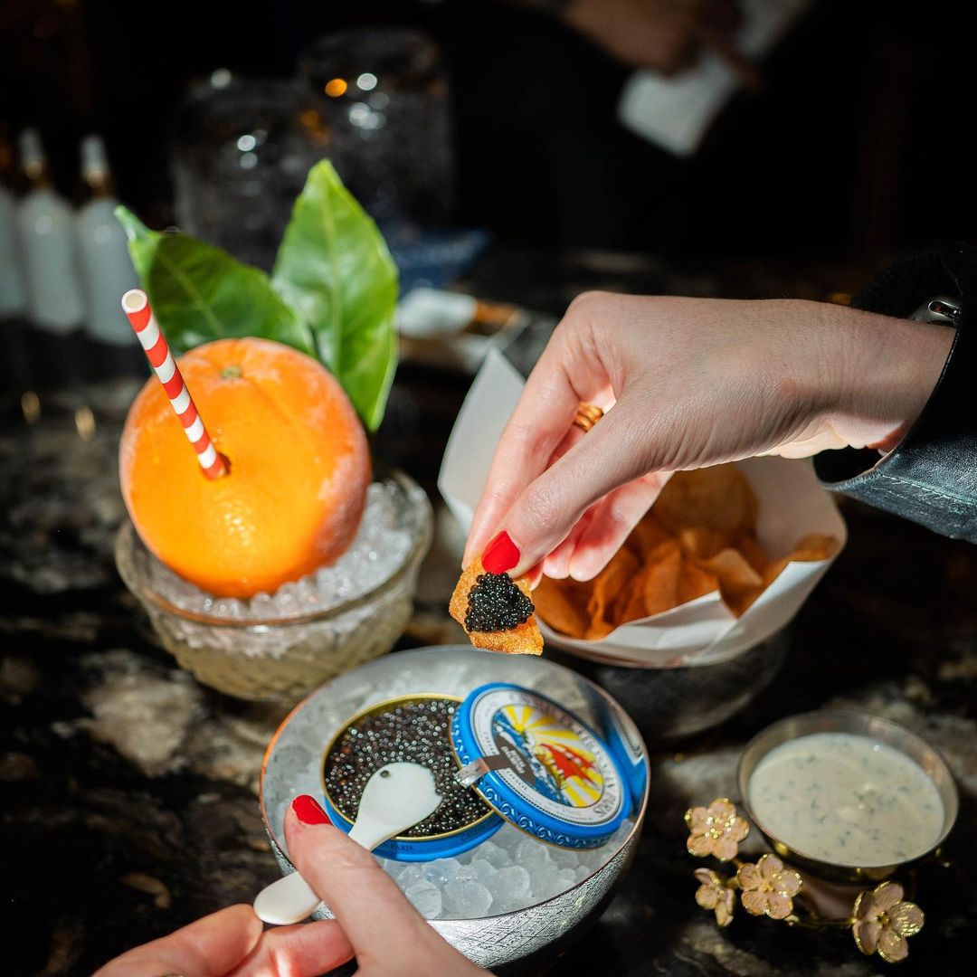 Close-up of a person's hands holding a toast with caviar, beside an orange drink with a straw and a bowl of caviar and chips.
