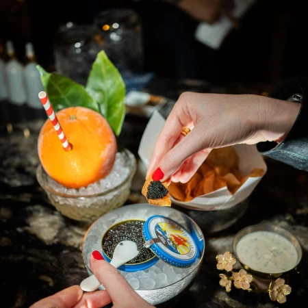Close-up of a person's hands holding a toast with caviar, beside an orange drink with a straw and a bowl of caviar and chips.