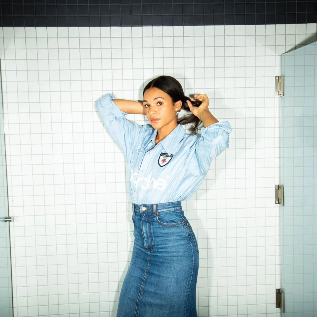 Young woman poses in blue top and denim skirt against a tiled wall, holding her hair with both hands in a casual indoor shot.