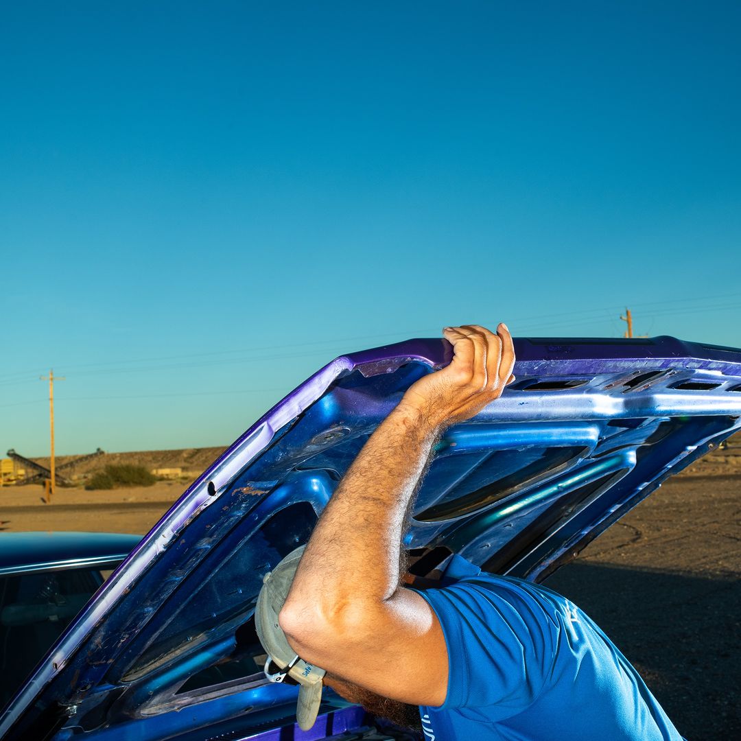 Man in blue shirt works on a car engine outdoors with the hood up against a clear blue sky. The car is blue and purple.