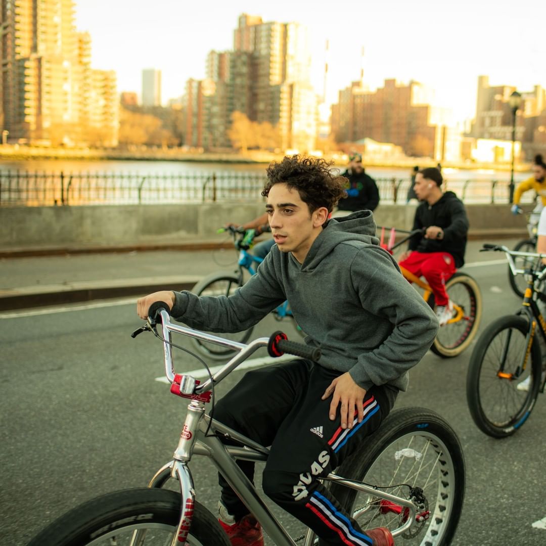 Young man riding a bicycle on a city street with buildings and river in the background, part of a group ride, wearing a hoodie.