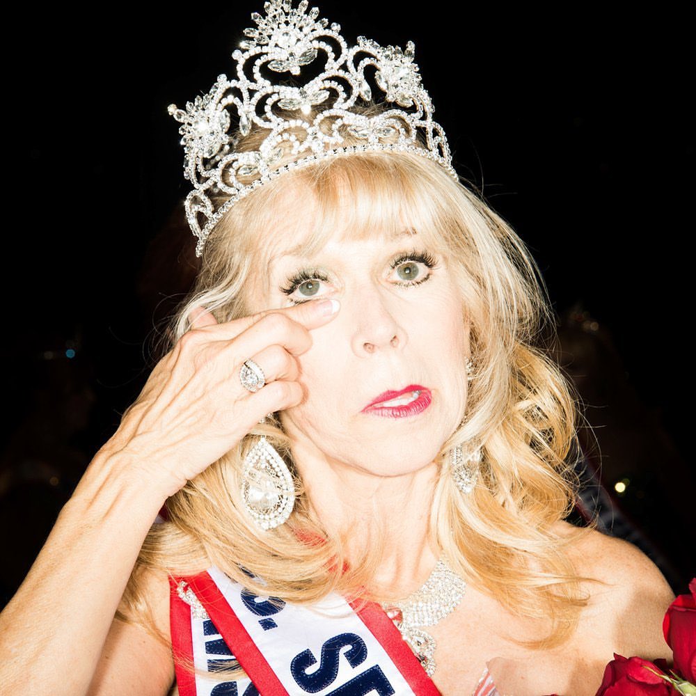 Mature woman crowned in tiara, wearing a sash and jewelry, touches eye. Shot against a black background, holding red roses.