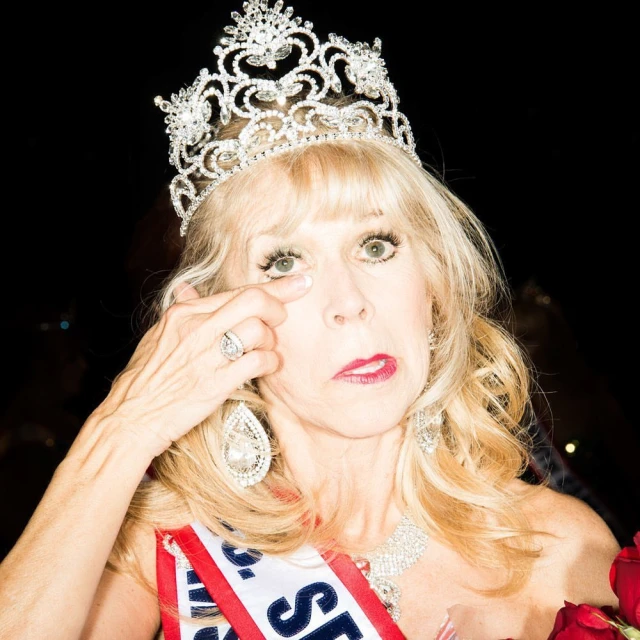 Mature woman crowned in tiara, wearing a sash and jewelry, touches eye. Shot against a black background, holding red roses.