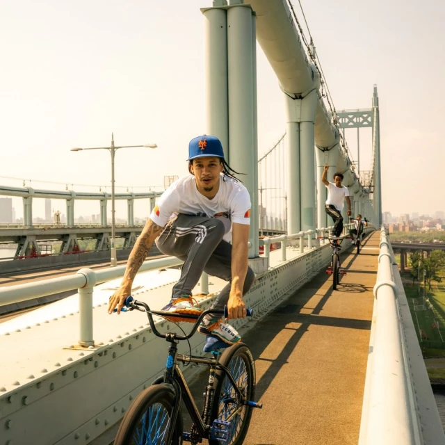 A man does stunts on a bike, balancing on the handlebars on a bridge with a city skyline in the background on a sunny day.