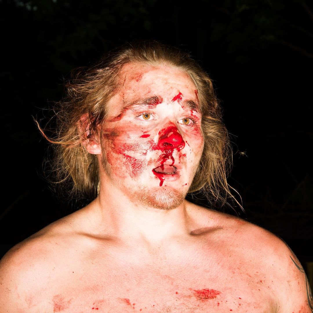 Dramatic close-up of adult male with fake blood on face and body, captured in dark outdoor environment with dramatic lighting.