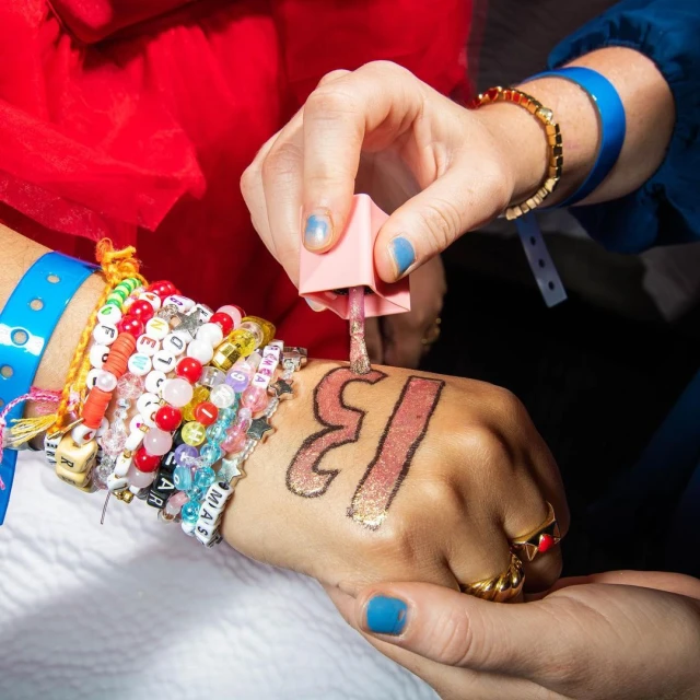 Close-up of an adult hand getting nail polish applied over a drawn-on design with colorful beaded bracelets visible.