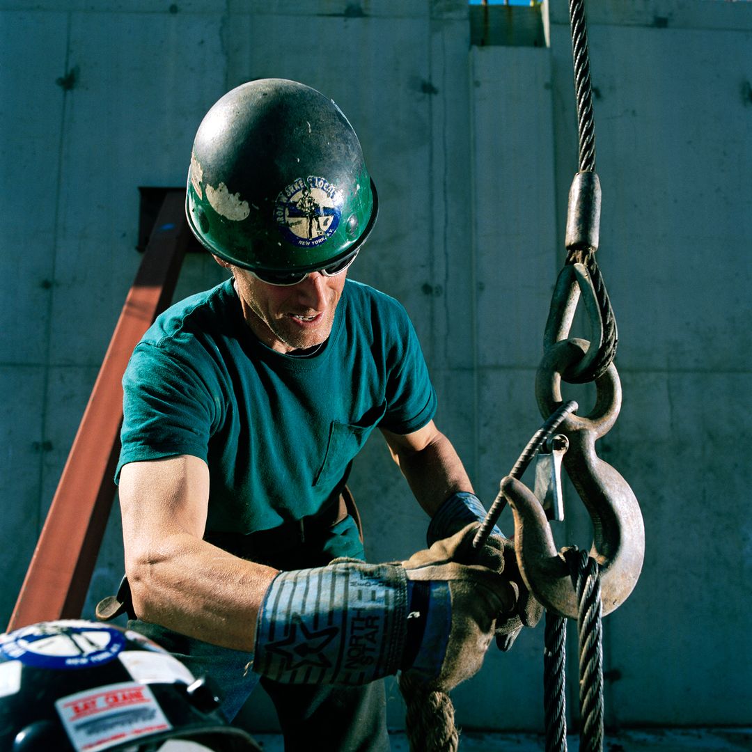 Man in a hardhat working with heavy-duty hooks and rigging equipment at a construction site, with a ladder visible in the background.