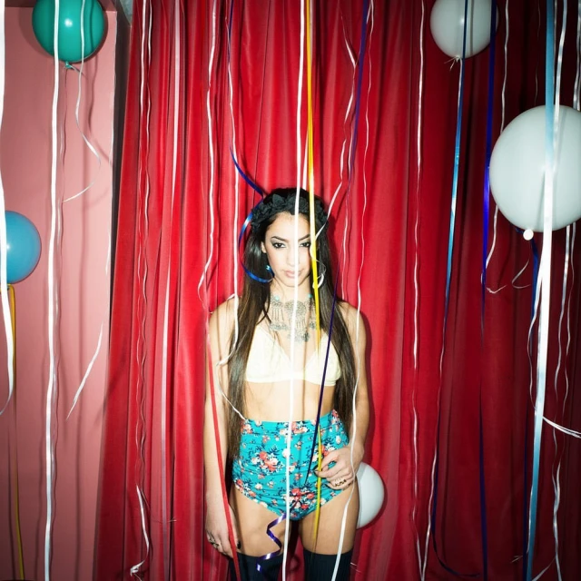 Woman in floral shorts stands in front of a red curtain with balloons and streamers, creating a festive, bright, and colorful scene.