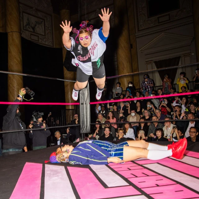 Two people in a wrestling ring. One person is in mid-air, seemingly jumping or diving toward the other, who is lying on the ring floor.