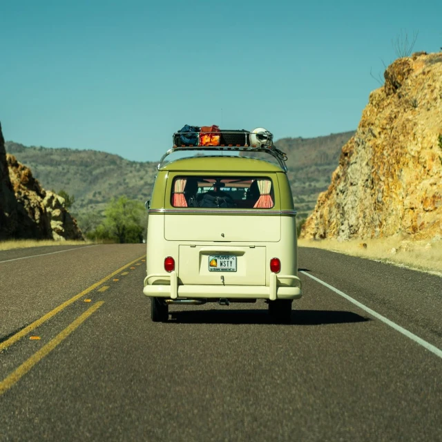 Rear view of a vintage green and white Volkswagen bus driving on an asphalt road through a desert landscape with rocky hills.