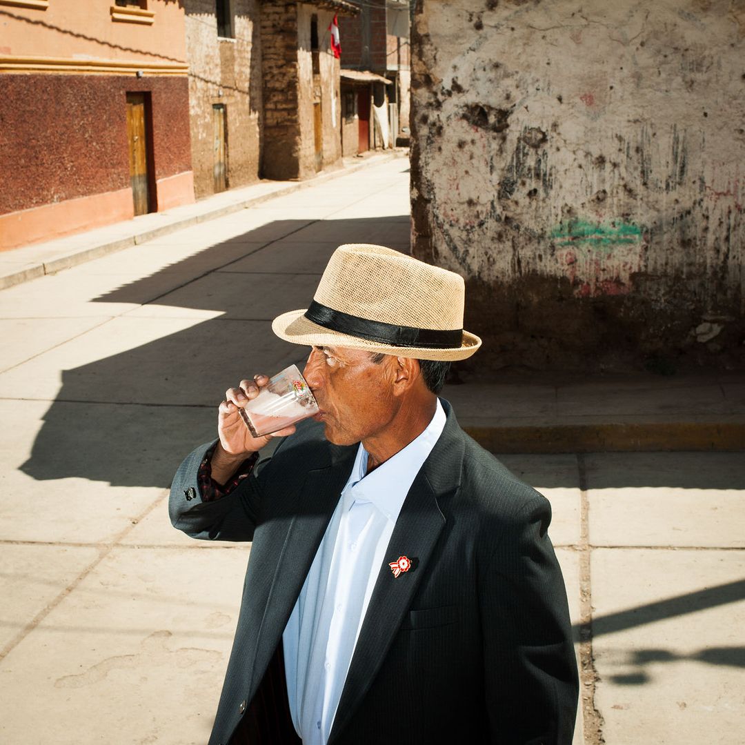 Adult man in a suit and hat drinks from glass on street. The old town has buildings with varied textures and colors in the background.