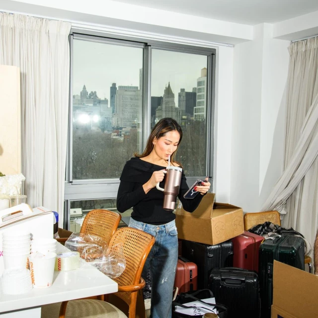 Woman in apartment drinking from a large cup, using a cell phone, with boxes and city views in the background, indoor, eye-level angle.