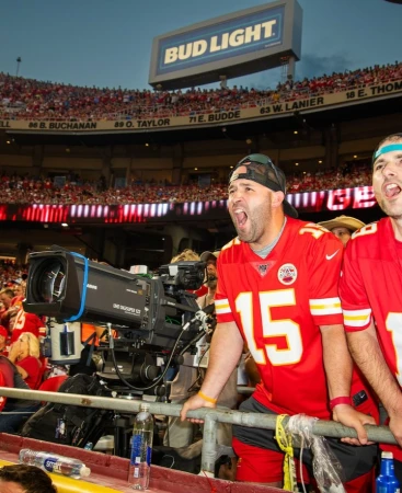 Fans cheer at a football game with a TV camera in the foreground and stadium seating with spectators in the background at night.