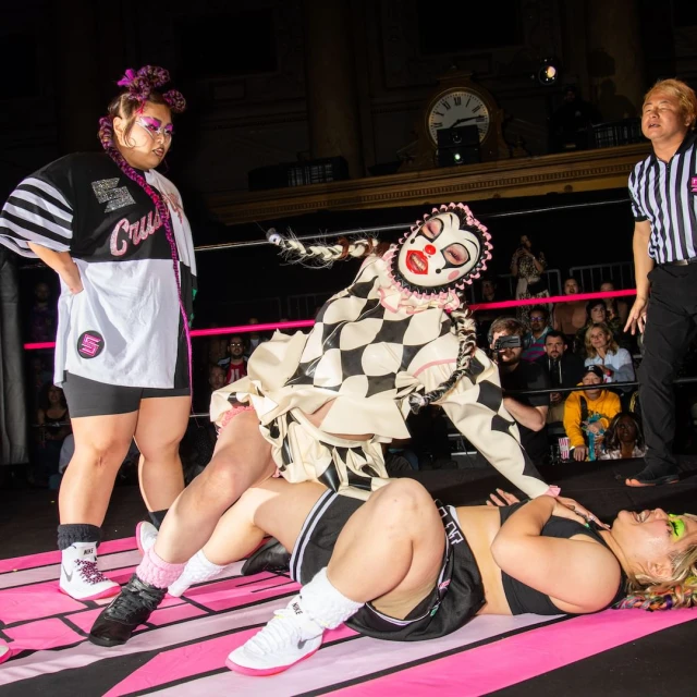 A wrestling match with a clown character pinning an opponent while a referee and another wrestler stand nearby in an indoor arena.