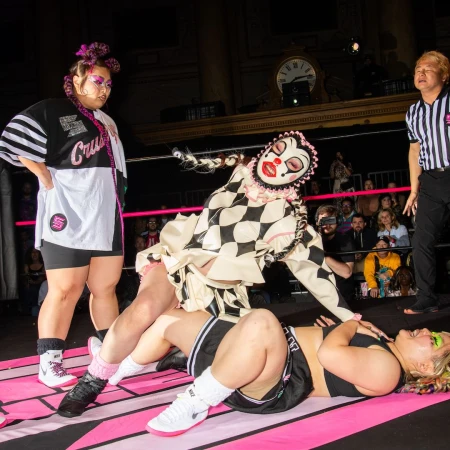 A wrestling match with a clown character pinning an opponent while a referee and another wrestler stand nearby in an indoor arena.