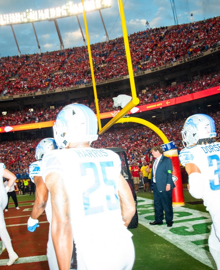 American football players in white and blue uniforms on a field with a large crowd in the background, football goalposts are visible.